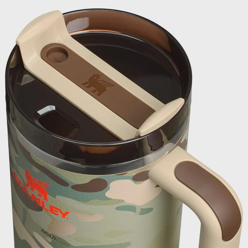 May include: A camouflage-patterned travel mug with a tan and brown lid and handle. The mug features the Stanley logo in red and the word "MULTICAM" in white. The lid has a brown accent with the Stanley logo embossed.