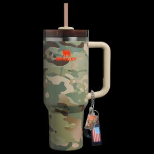 May include: A camouflage-patterned Stanley tumbler with a handle, straw, and keychain. The tumbler features the Stanley logo in orange. The handle and straw are beige. The keychain has a small American flag.