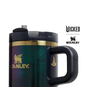 Op de afbeelding: Een zwarte en iriserende Stanley-mok met een handvat, met het "Wicked"-logo en het Stanley-logo. De mok heeft een gouden band en een zwart deksel. Het Stanley-logo is een gevleugelde beer. De mok is ontworpen om dranken warm of koud te houden.