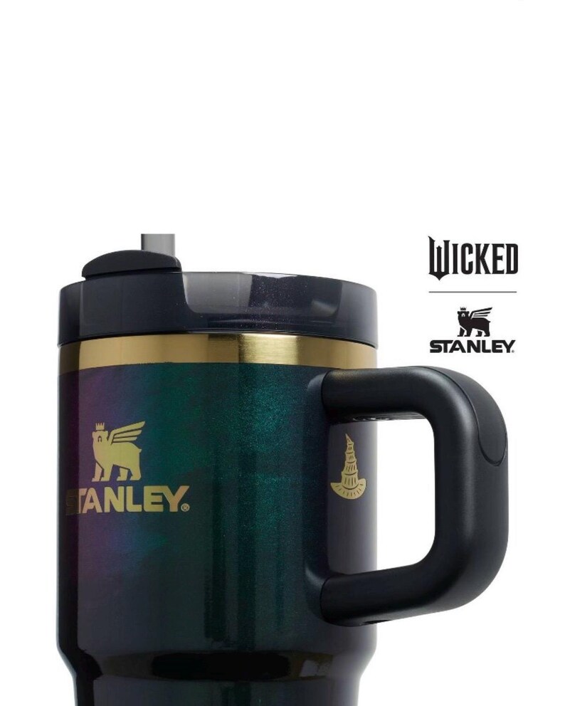 Op de afbeelding: Een zwarte en iriserende Stanley-mok met een handvat, met het "Wicked"-logo en het Stanley-logo. De mok heeft een gouden band en een zwart deksel. Het Stanley-logo is een gevleugelde beer. De mok is ontworpen om dranken warm of koud te houden.
