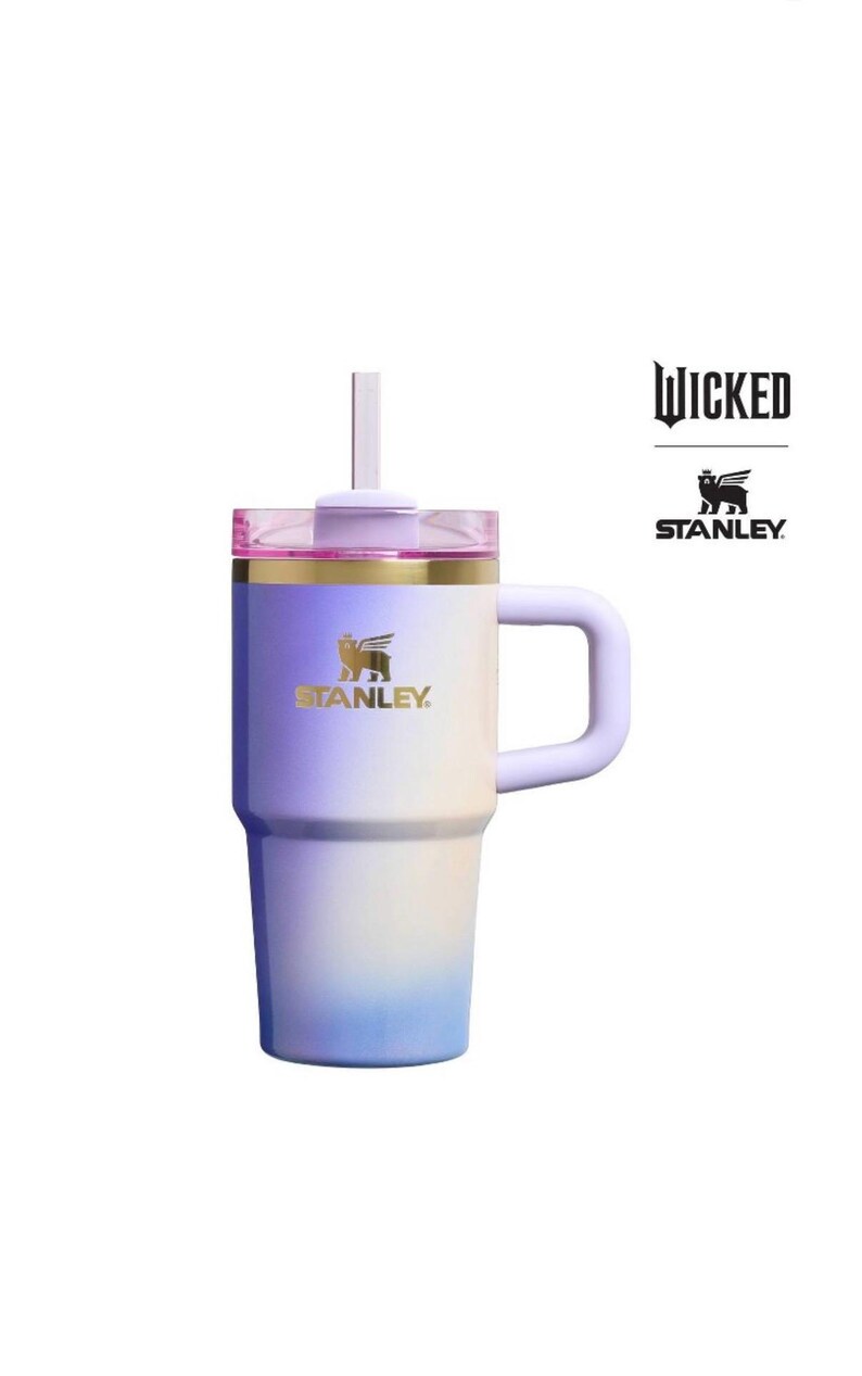 Op de afbeelding: Een Stanley-beker met een kleurovergang in paars, wit en blauw. Hij heeft een handvat, een deksel met een rietje en het Stanley-logo in goud. Het woord "Wicked" staat rechtsboven gedrukt.