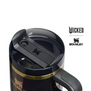 Op de afbeelding: Zwarte Stanley reismok met een gouden band en handvat. De mok heeft de logo's "Wicked" en "Stanley". De deksel is zwart met een schuifsluiting en een klein reli&euml;flogo. Ontworpen voor warme of koude dranken.
