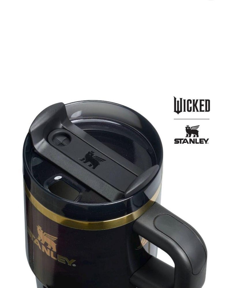 Op de afbeelding: Zwarte Stanley reismok met een gouden band en handvat. De mok heeft de logo's "Wicked" en "Stanley". De deksel is zwart met een schuifsluiting en een klein reli&euml;flogo. Ontworpen voor warme of koude dranken.