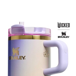 Op de afbeelding: Een Stanley-mok met een gradi&euml;ntontwerp in tinten paars, goud en wit. De mok heeft een handvat en een deksel met een rietje. Het Stanley-logo en het woord "Wicked" zijn zichtbaar.