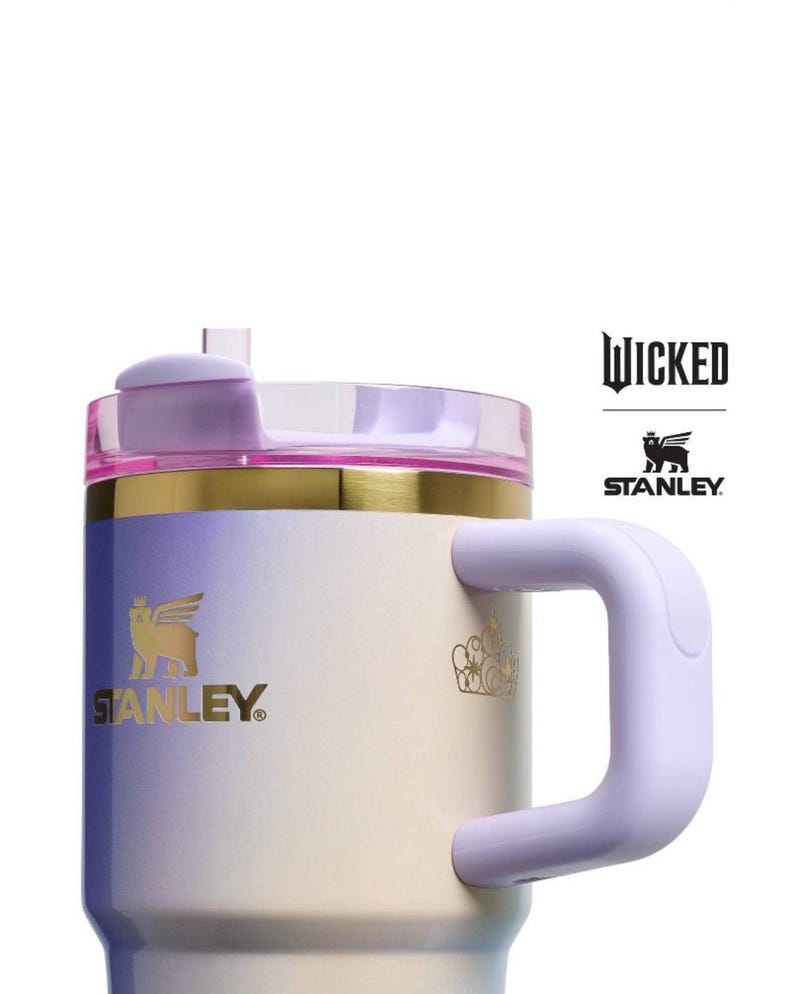 Op de afbeelding: Een Stanley-mok met een gradi&euml;ntontwerp in tinten paars, goud en wit. De mok heeft een handvat en een deksel met een rietje. Het Stanley-logo en het woord "Wicked" zijn zichtbaar.