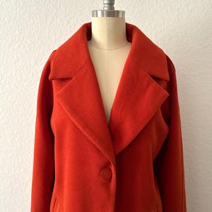 May include: A long, red, collared coat with a single button closure. The coat has a slightly oversized fit and a relaxed silhouette.