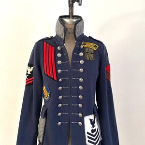 May include: A navy blue military-style jacket featuring a high collar with grey herringbone fabric, silver-toned buttons, and various embroidered patches. The jacket includes red stripes, a Comando Motorizado patch, and eagle insignias.
