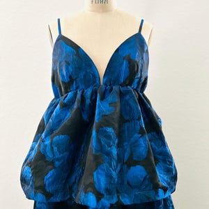 May include: A blue and black floral print dress with a deep V-neckline and spaghetti straps. The dress features a voluminous, layered design with a textured fabric, creating a dramatic silhouette. The dress is displayed on a mannequin.