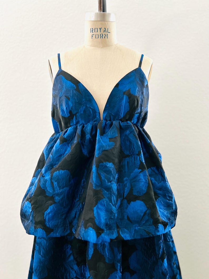 May include: A blue and black floral print dress with a deep V-neckline and spaghetti straps. The dress features a voluminous, layered design with a textured fabric, creating a dramatic silhouette. The dress is displayed on a mannequin.