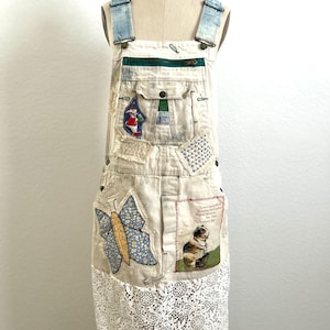 May include: Off-white denim overalls featuring light blue denim straps, a green zippered chest pocket, and a Liberty brand patch. The garment is decorated with a blue floral butterfly appliqué, a small embroidered cat patch, and a white lace skirt.