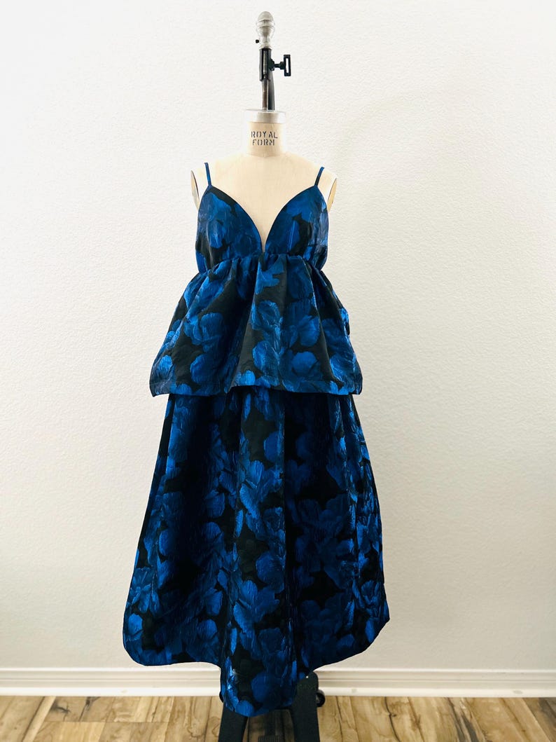 May include: A two-piece formal dress with a deep V-neckline and spaghetti straps. The dress features a blue floral pattern on a black background. The top has a peplum design, and the skirt is a midi length.