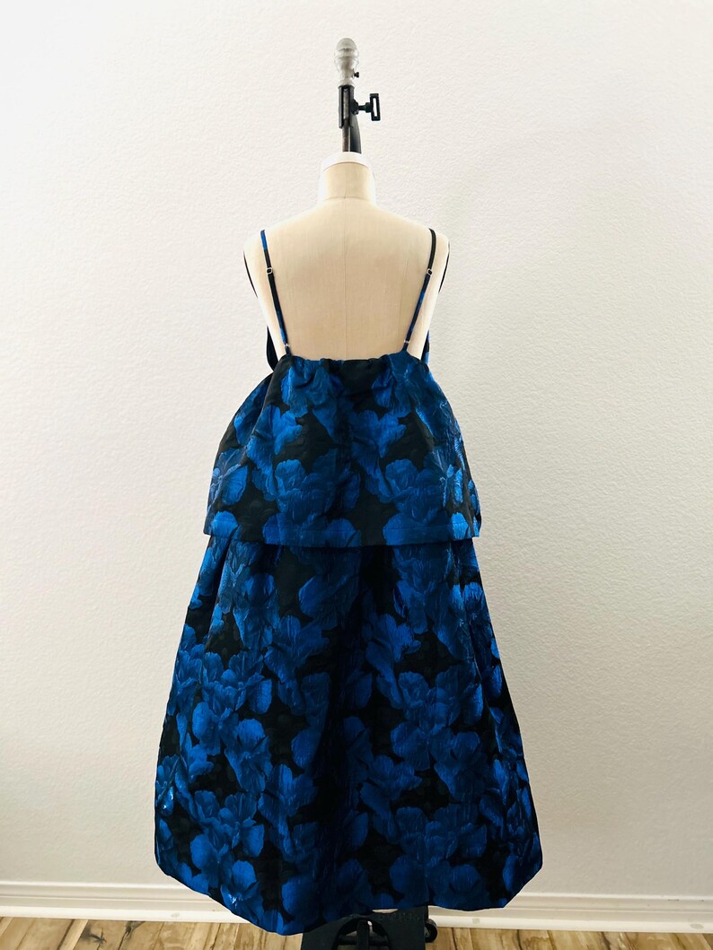 May include: A formal dress with a blue floral pattern on a black background. The dress features a layered design with spaghetti straps. The garment is displayed on a mannequin against a white backdrop.