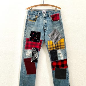 May include: Light blue denim jeans with various fabric patches in red, black, yellow, and white. The patches are in plaid, floral, and polka dot patterns. The jeans have a button fly and are distressed with rips.