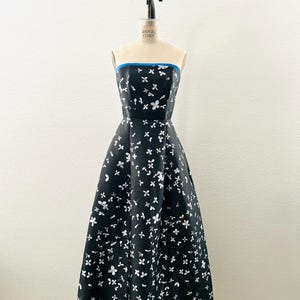 May include: A strapless black dress with a white floral pattern and a blue trim. The dress is displayed on a mannequin. The skirt flares out from the waist and has a blue lining visible at the hem.