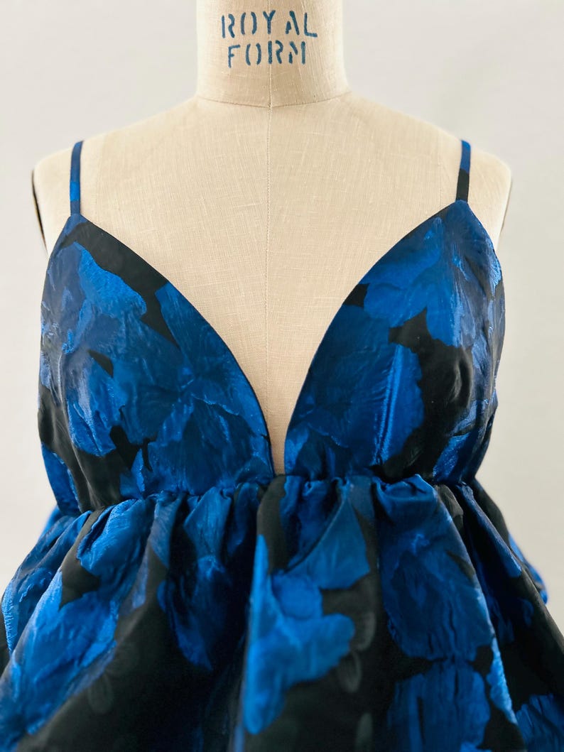 May include: A sleeveless top with a deep V-neck and a floral pattern in shades of blue and black. The top is displayed on a mannequin with the text "ROYAL FORM" at the top. The fabric has a textured appearance.