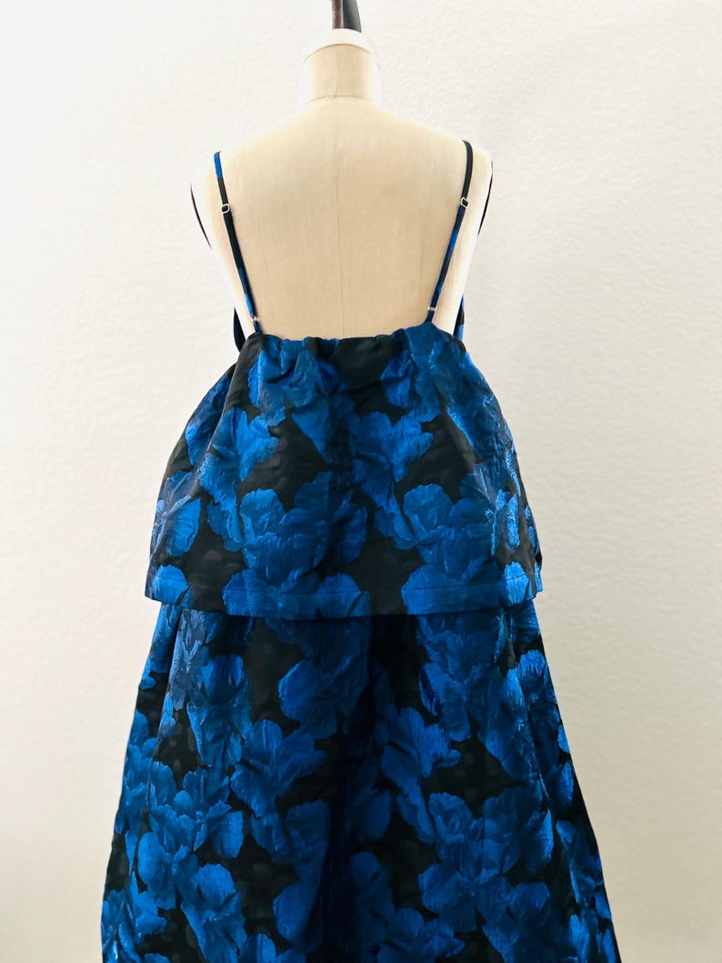 May include: A two-piece formal dress with a black base and a vibrant blue floral pattern. The top features thin straps and a layered design, while the skirt flows down. The dress is displayed on a mannequin.