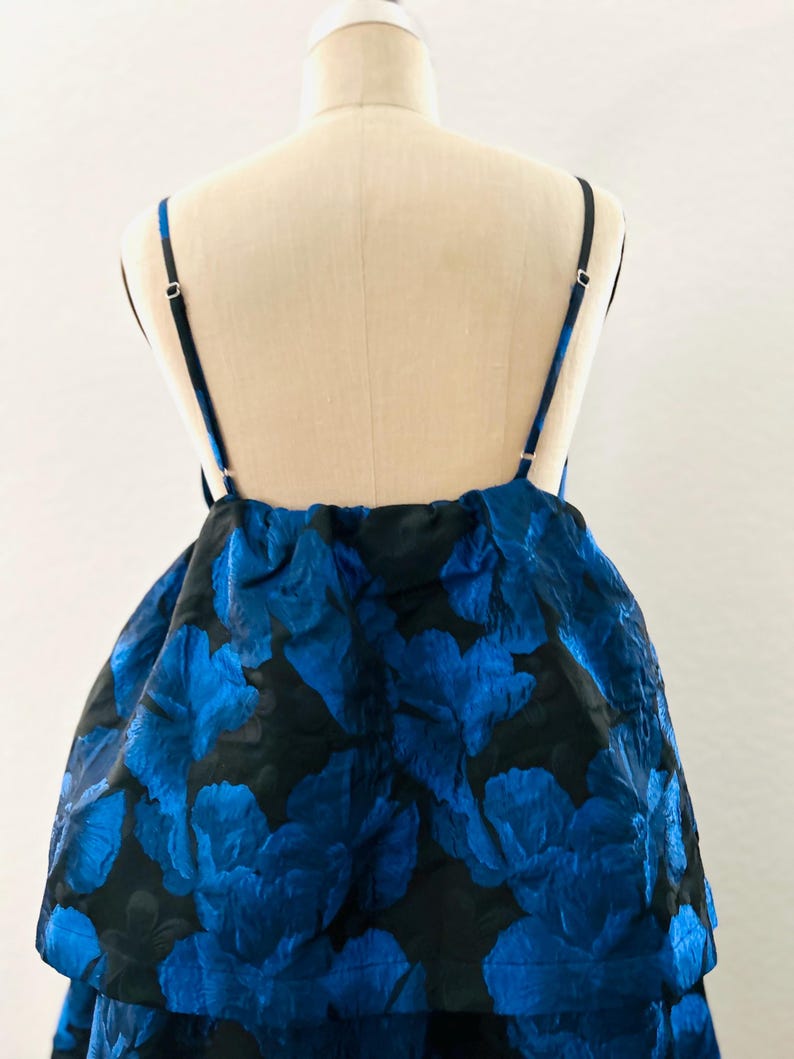 May include: A blue and black floral print dress on a mannequin. The dress features a layered skirt and thin black straps. The floral pattern is a vibrant blue against a black background, creating a striking contrast. The dress is designed for a formal occasion.