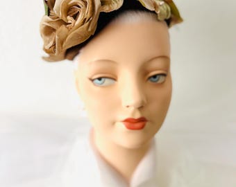 May include: A vintage hat featuring large, fabric roses in shades of tan and brown, accented with green leaves. The floral arrangement sits atop a dark band. The hat is displayed on a mannequin head with blue eyes and red lipstick.