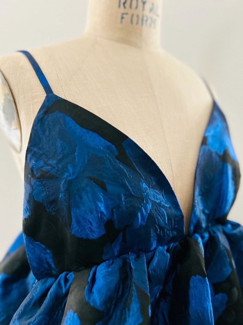 May include: A sleeveless dress with a deep V-neckline. The fabric features a bold floral pattern in shades of blue and black. The dress is displayed on a mannequin, showcasing its design and fit. The dress has thin straps.