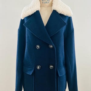 May include: Navy blue double-breasted coat with a cream-colored shearling collar. The coat features two front pockets and four dark buttons. The coat is displayed on a mannequin.