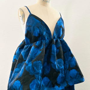 May include: A blue floral print dress with a deep V-neck and spaghetti straps. The dress features a voluminous skirt with a layered design. The fabric has a textured appearance, and the dress is displayed on a mannequin.