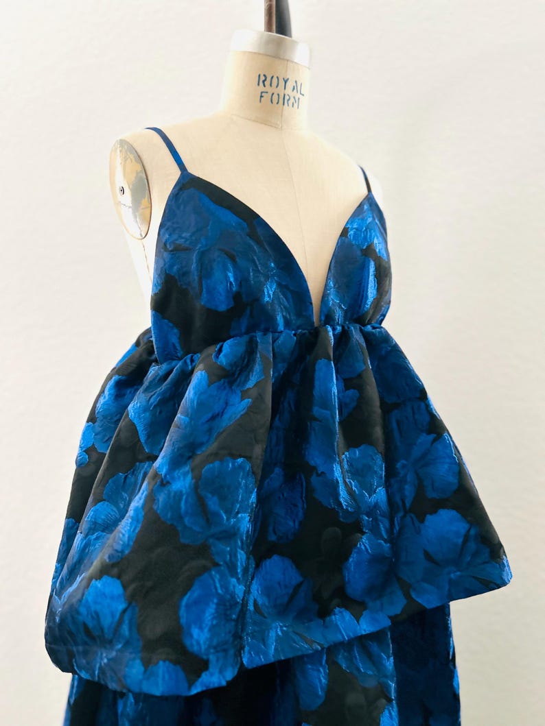 May include: A blue floral print dress with a deep V-neck and spaghetti straps. The dress features a voluminous skirt with a layered design. The fabric has a textured appearance, and the dress is displayed on a mannequin.