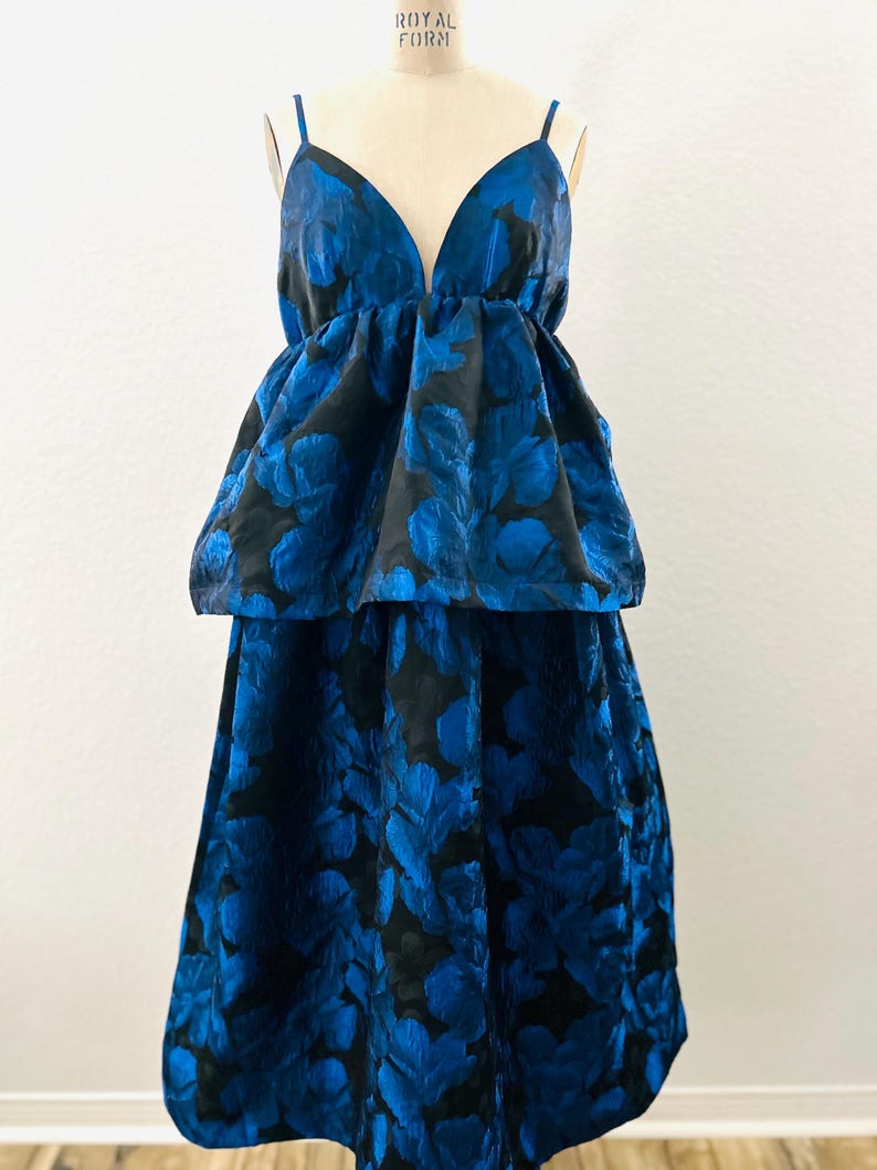 May include: A two-piece formal dress with a deep V-neckline and spaghetti straps. The dress features a black background with a vibrant blue floral pattern. The top has a peplum design, and the skirt is a midi length.