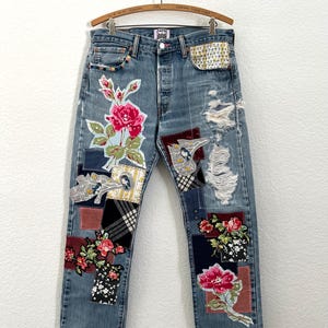 May include: Light blue denim jeans with patches and floral embroidery. The jeans feature a distressed design with ripped areas and various fabric patches. Patches include floral patterns, plaid, and other decorative elements. The jeans are hanging on a wooden hanger.