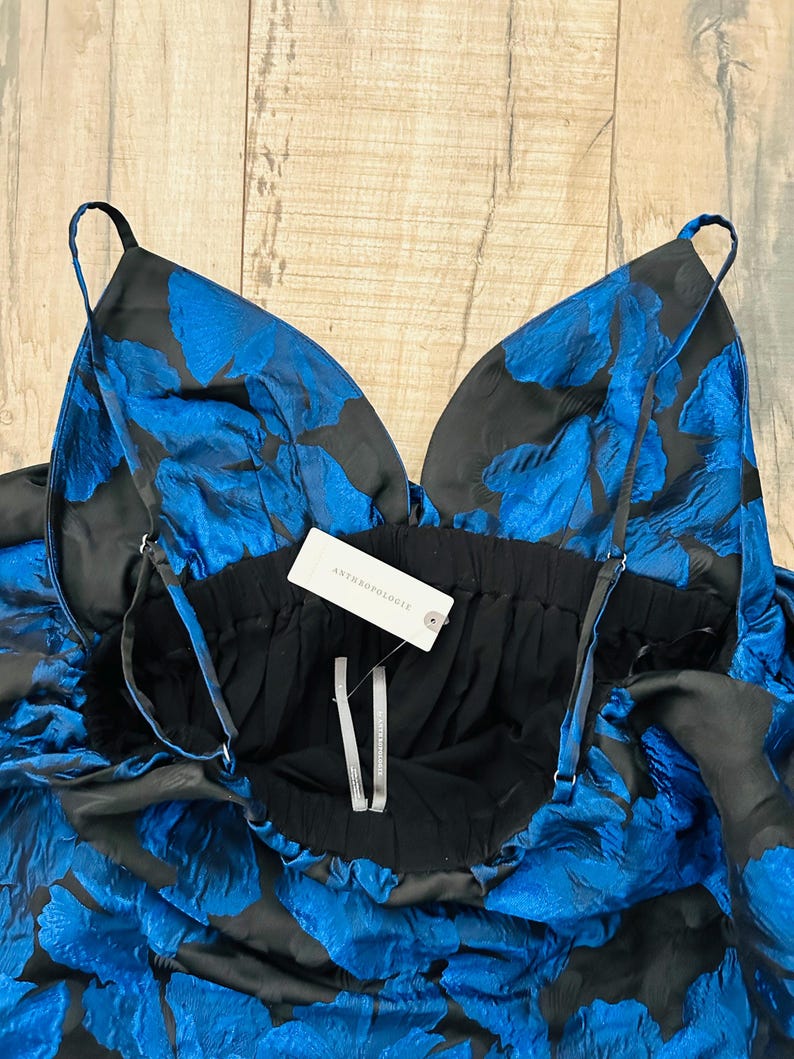 May include: A black and blue floral print dress with thin straps. The dress features a deep V-neckline and a gathered waist. The fabric has a textured appearance. A tag that says "Anthropologie" is attached to the dress.