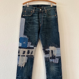 May include: Dark wash denim jeans with patchwork detailing in various shades of blue and white. The jeans are hanging on a wooden hanger. The patchwork is on the front of the legs.