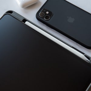Peut inclure: Une coque noire pour iPad avec un stylet blanc reposant sur l'écran. La coque a une découpe pour l'Apple Pencil.