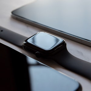 May include: A black Apple Watch with a black band on a white surface. The watch face is black and reflective.