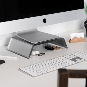 Peut inclure: Un bureau blanc avec un écran d'ordinateur argenté sur un support argenté. Un clavier sans fil blanc est sur le bureau. Une tablette noire et un stylet blanc sont sur le bureau. Un porte-cartes de visite en bois est sur le bureau. Un casque noir est sur le bureau.
