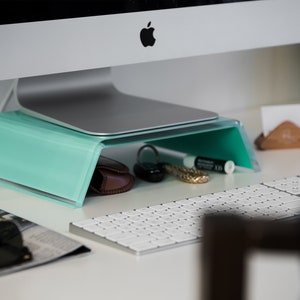 Peut inclure: Un support d'écran en acrylique bleu turquoise avec un écran d'ordinateur argenté sur le dessus. Le support est sur un bureau blanc avec un clavier blanc, un magazine, des lunettes de soleil et un portefeuille en cuir marron en dessous.