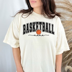 May include: A cream colored t-shirt with black text that reads "Basketball mama" and a black and orange basketball graphic.