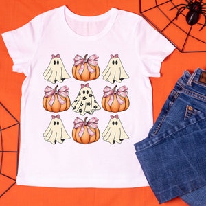 May include: White t-shirt featuring a Halloween-themed design. The design includes cartoon ghosts with pink bows and pumpkins with pink bows. The shirt is paired with blue jeans on an orange background.