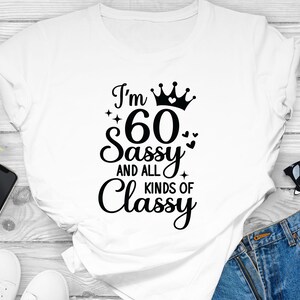 以下が含まれることがあります： 黒い文字で「I'm 60 Sassy and all kinds of Classy」と書かれた白いTシャツ。文字の上に王冠とハートが描かれている。
