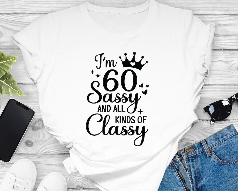 以下が含まれることがあります： 黒い文字で「I'm 60 Sassy and all kinds of Classy」と書かれた白いTシャツ。文字の上に王冠とハートが描かれている。
