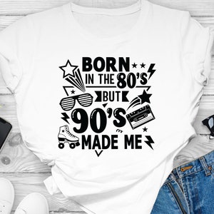 May include: White t-shirt with a black graphic design that says "Born in the 80's but 90's made me". The graphic includes a star, a lightning bolt, roller skates, sunglasses, and a cassette tape.