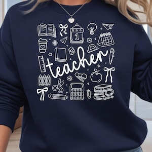 May include: Navy blue sweatshirt with a white graphic design featuring school-related icons and the word "teacher". The design includes books, a coffee cup, a calendar, a lightbulb, and other school supplies.