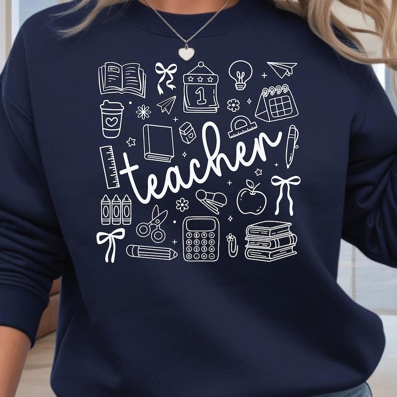 May include: Navy blue sweatshirt with a white graphic design featuring school-related icons and the word "teacher". The design includes books, a coffee cup, a calendar, a lightbulb, and other school supplies.