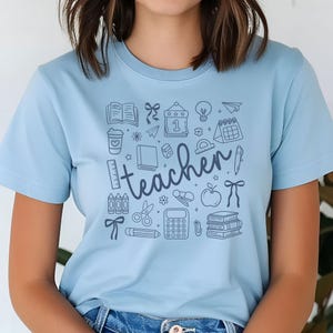 May include: Light blue t-shirt with a teacher-themed graphic. The design includes school-related icons such as books, pencils, an apple, and the word "teacher" in a handwritten font.
