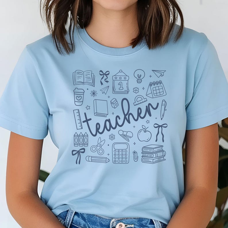 May include: Light blue t-shirt with a teacher-themed graphic. The design includes school-related icons such as books, pencils, an apple, and the word "teacher" in a handwritten font.
