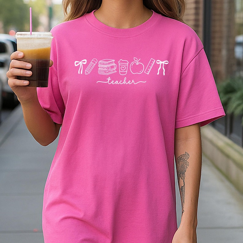 May include: A bright pink t-shirt with a white graphic of school supplies and the word "teacher" in a cursive font. The graphic includes a pencil, books, a coffee cup, an apple, and a ruler. The shirt is a short-sleeved, casual style.