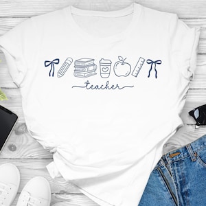 May include: White t-shirt with a navy blue graphic featuring school-related icons such as a pencil, books, coffee cup, apple, ruler, and a bow. The word "teacher" is written in cursive below the icons.