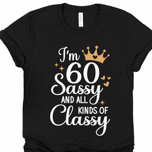 以下が含まれることがあります： 黒いTシャツに白い文字で「I'm 60 Sassy and all kinds of Classy」と書かれています。文字は金の王冠とハートで飾られています。