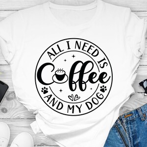 May include: White t-shirt with a black circle graphic that says "All I need is coffee and my dog" with a coffee cup, paw prints, and hearts.