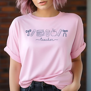 May include: A light pink t-shirt with rolled-up sleeves. The shirt features a dark blue graphic of school-related items, including a pencil, books, a coffee cup, an apple, and a ruler. The word "teacher" is written in cursive below the graphics.