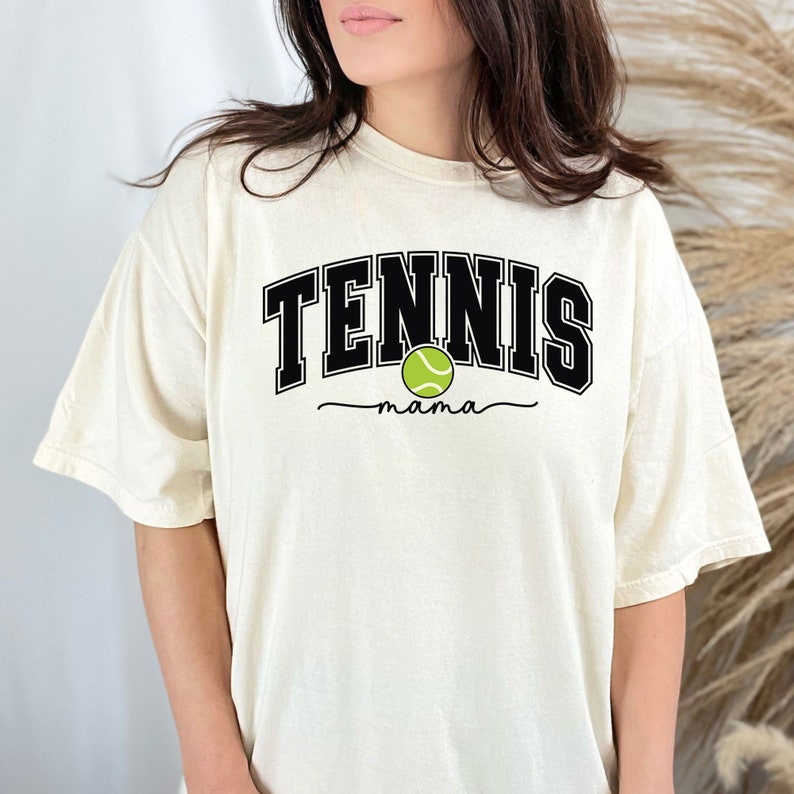 Może przedstawiać: Biała koszulka z czarnym napisem "TENNIS mama" i zielonym obrazkiem piłki tenisowej.