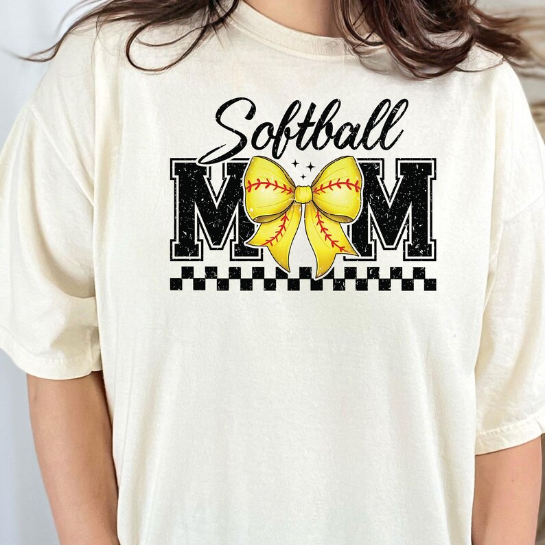 May include: A white t-shirt with black text that reads "Softball Mom" with a yellow softball bow in the middle.